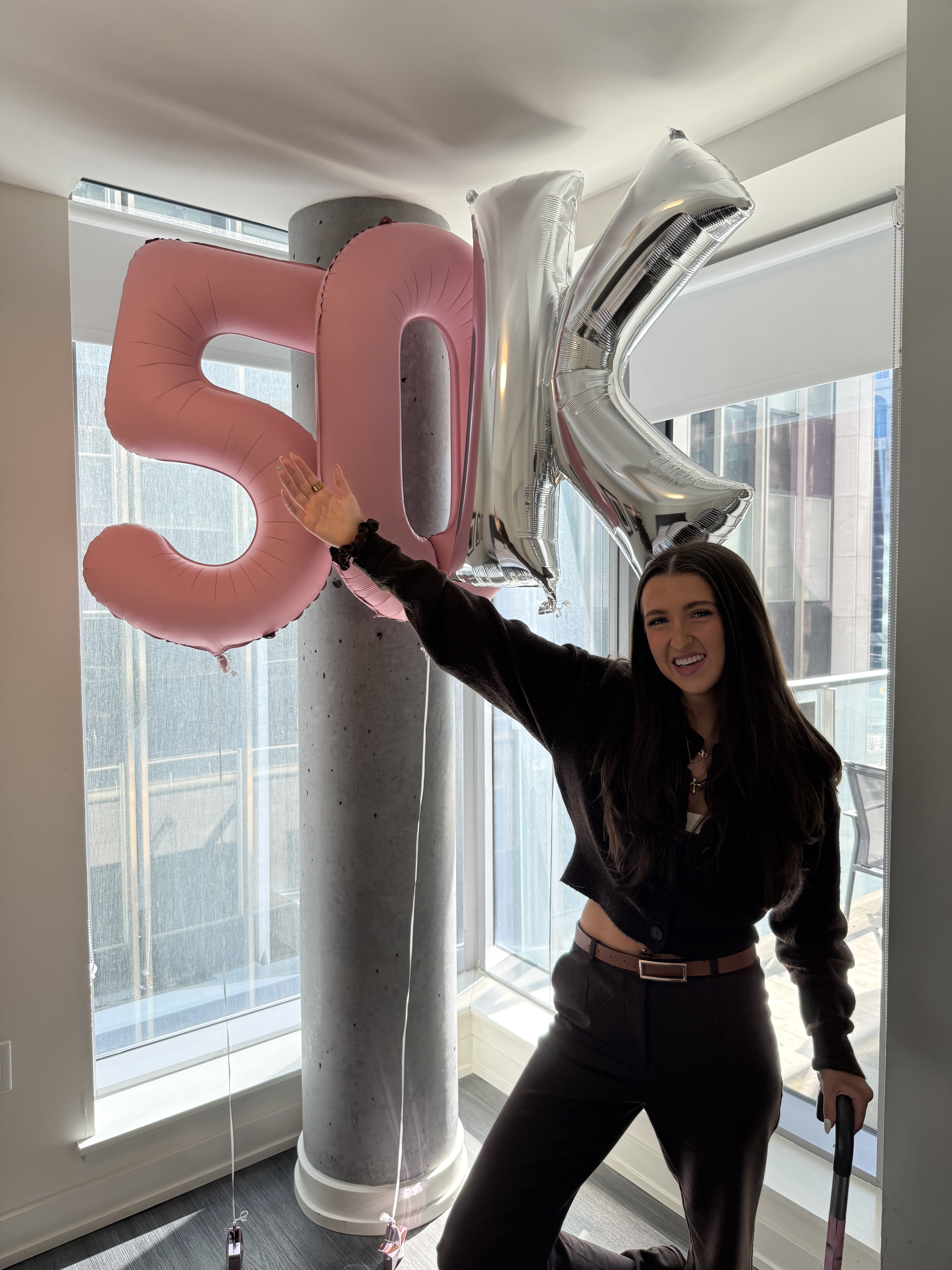 Kale Vine holding 50K balloon letters, smiling in a dark blazer with a cane, floor-to-ceiling windows behind her