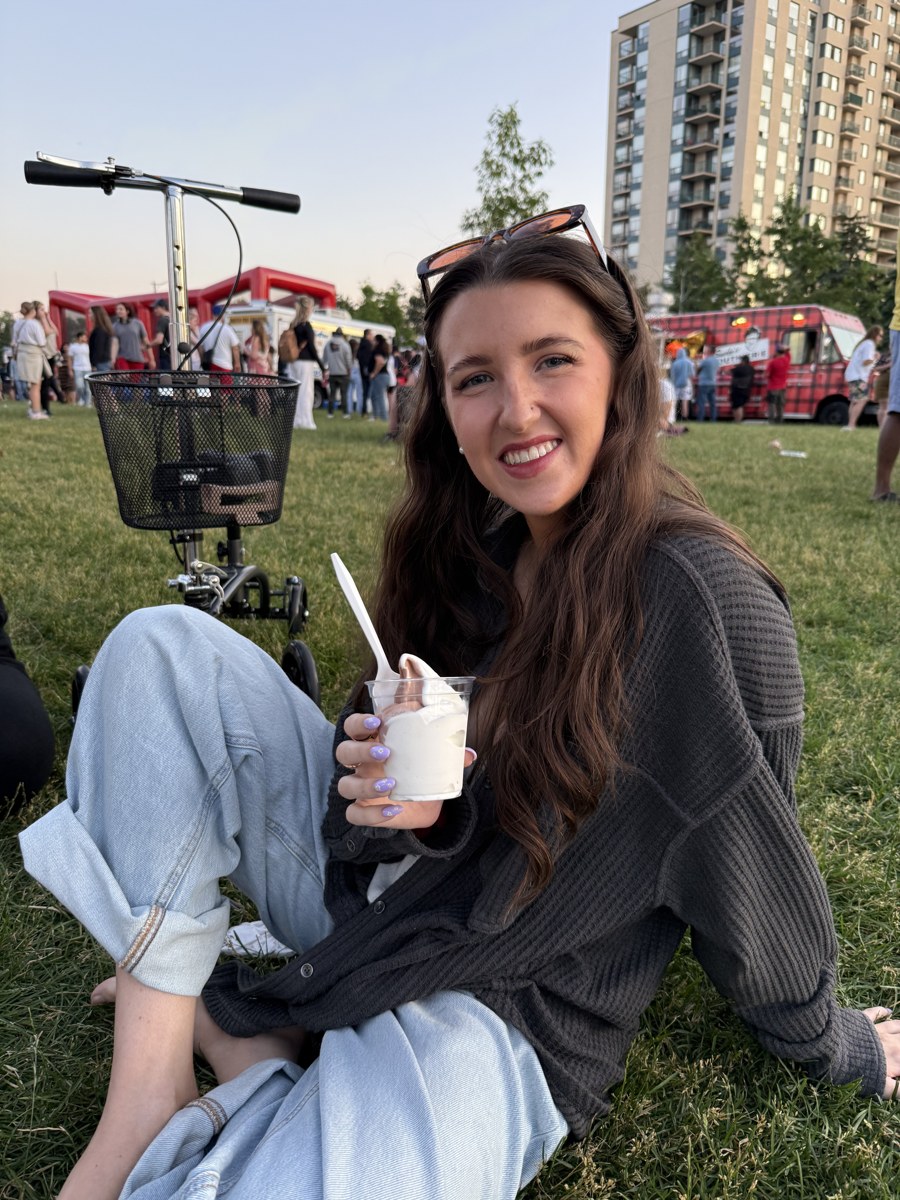 Kale Vine sitting on grass at an outdoor festival holding ice cream, knee scooter visible