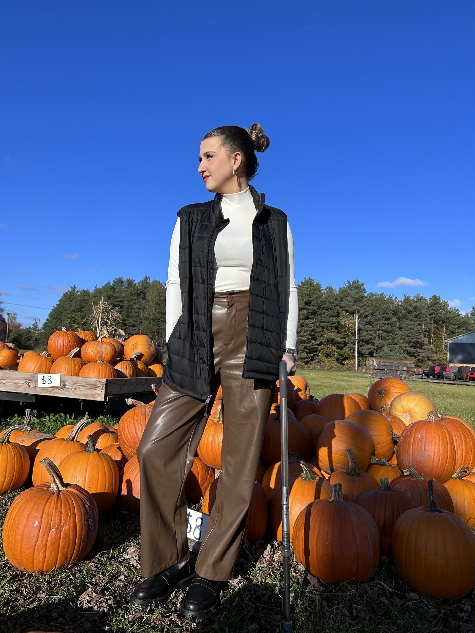 Kale Vine at a pumpkin patch in a white turtleneck and leather pants, holding a cane, blue sky